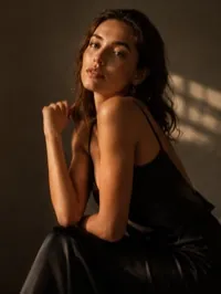 A model seated in a black slip dress under hard directional light