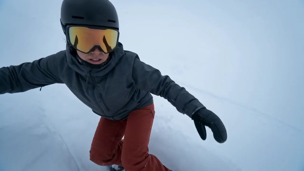 Red Bull spec action film — a snowboarder in a black helmet and rust-red pants carving through whiteout powder